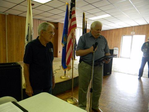 Two men standing at front of room, one with microphone and reading off a piece of paper