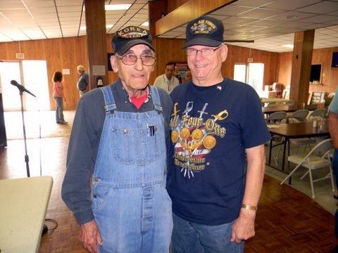 Man in overalls standing beside man in blue shirt, both are smiling