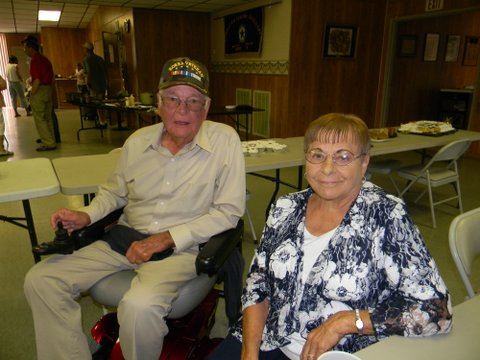 Man in motorized scooter beside woman in chair, both looking into camera smiling