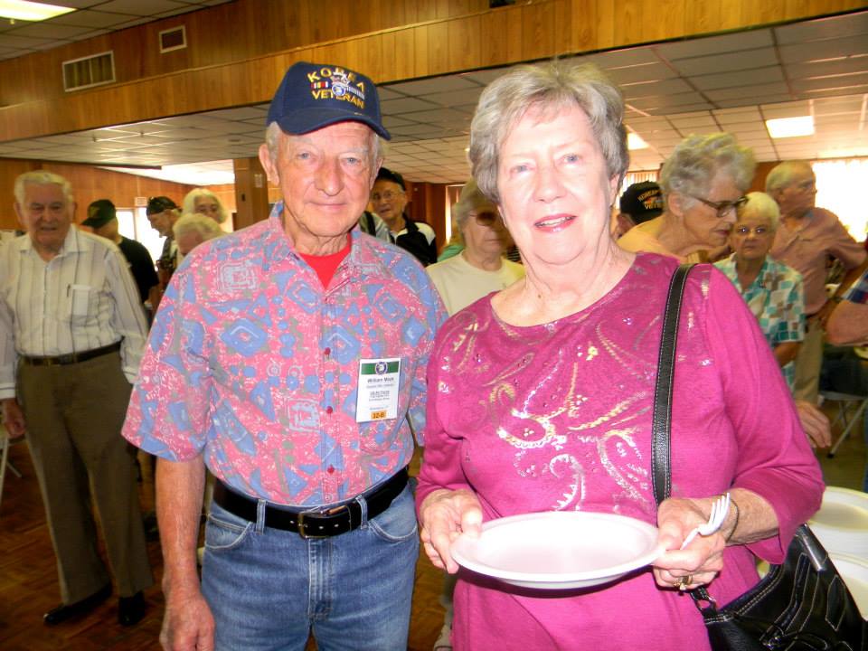 Man and woman, both in pink shirts, standing and smiling, woman is carrying a plate