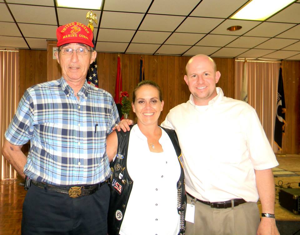 Man with red Korean war hat on, standing with woman wearing white an black, and a man with his arm around her shoulder