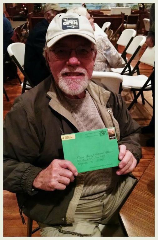 Sitting veteran with khaki hat on proudly hold up unopend green envelope, looking into the camera and smiling