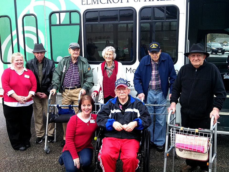 Two nurses stand with six veterans outside of bus in parking lot, smiling at camera