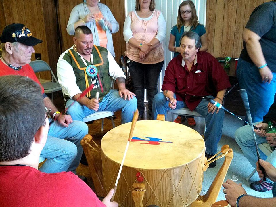 Man wearing Native American beads plays a large drum with others in circle