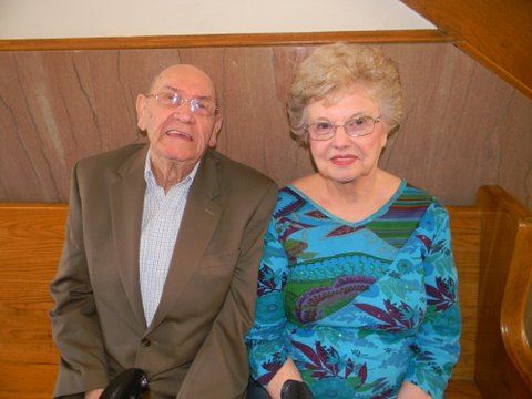 Man in khaki suit and woman in turquoise shirt sit on bench, smiling at camera