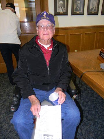 Man in motorized cart wears red shirt and black jacket as well as purple Purple Heart hat