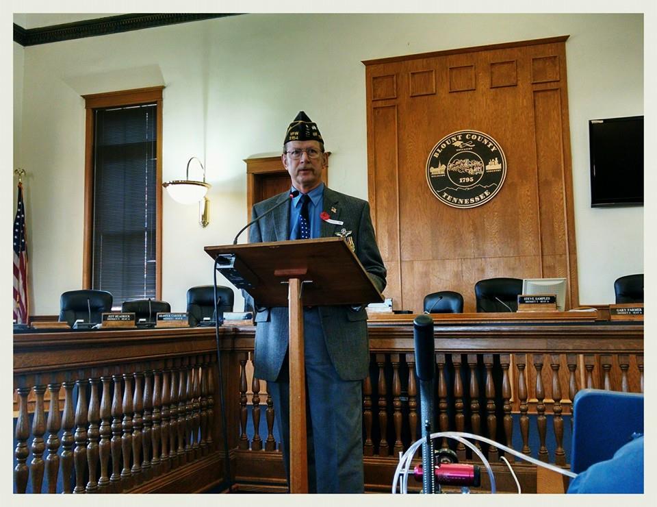 Man in grey suit and blue shirt and cap, speaks at wooden podium inside courtroom