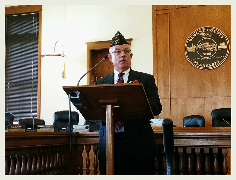Man in black suit and white shirt, glasses and cap, speaks at wooden podium inside courtroom