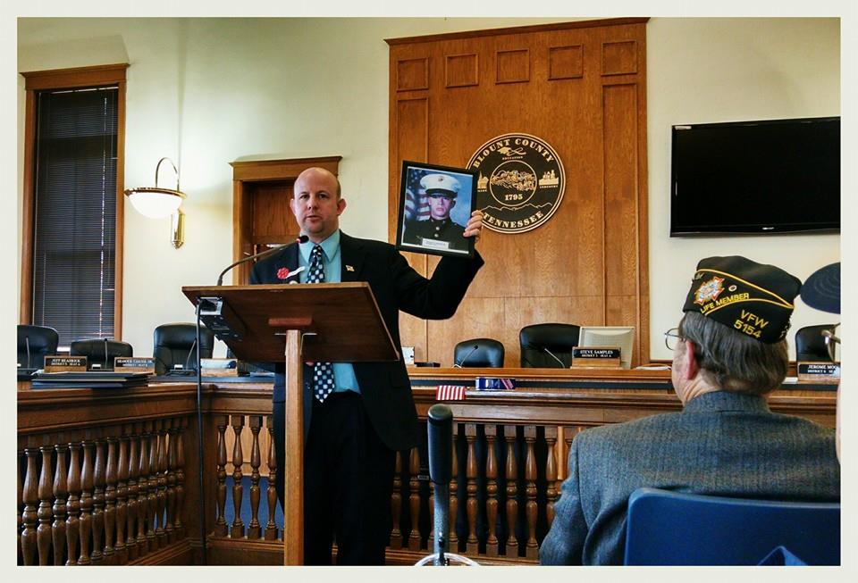 Man in black suit and blue shirt speaks at podium inside courtroom, holding up framed picture of marine