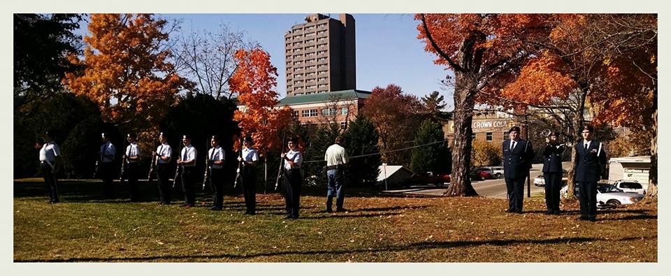 Eleven cadets stand in formation, one plays trumpet, seven with firearms in front of them