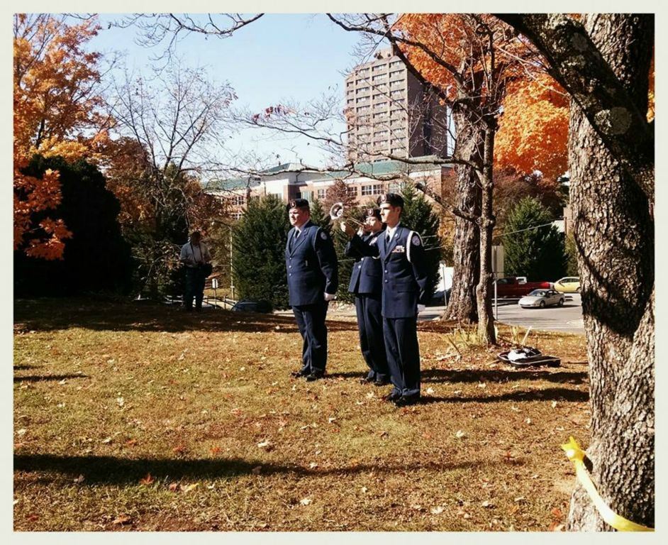 Three cadets, one playing trumpet, stand on lawn under trees