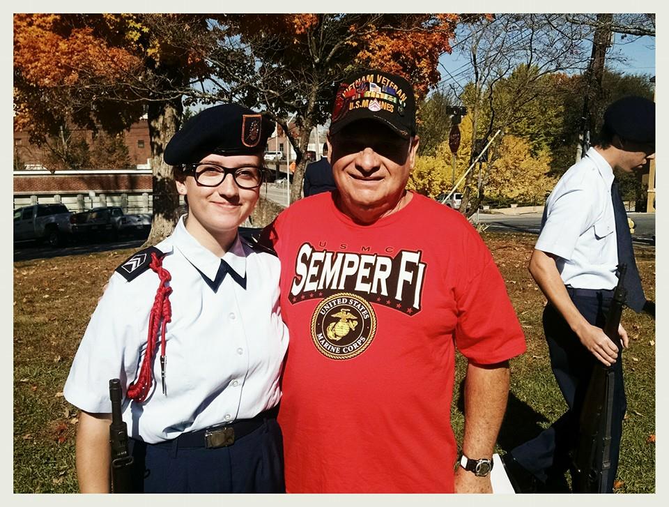 Young woman cadet stands smiling side hugging man in red Marine Corps shirt and Vietnam veteran hat
