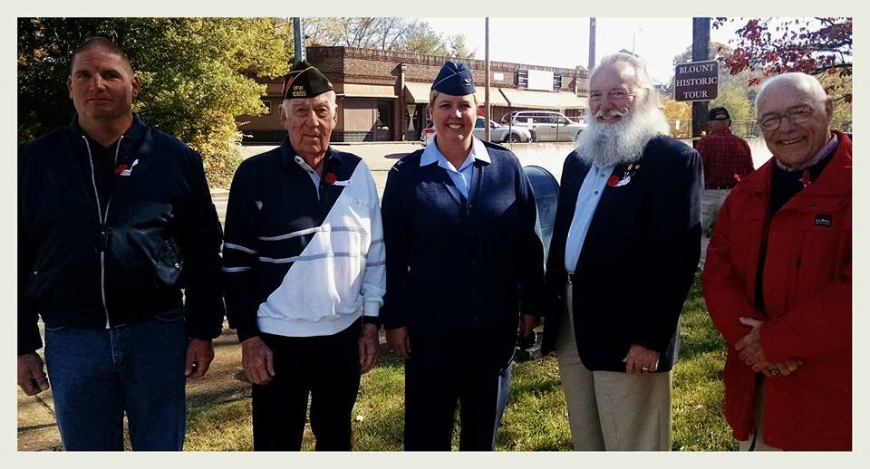 Five people, four men and one woman, stand in a line outside, smiling at camera
