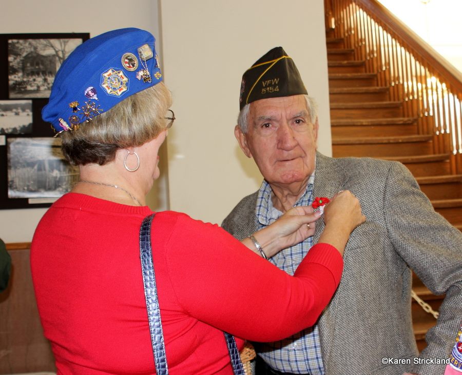 Lady in red shirt and blue hat pins poppy pin to veteran's grey lapel