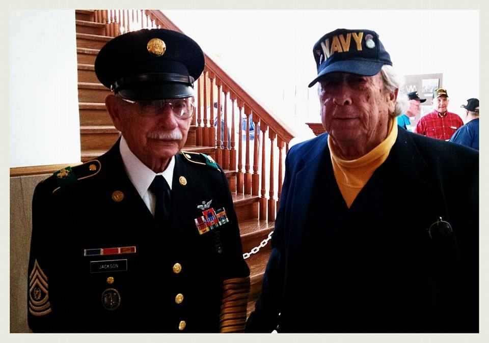 Man in dress uniform stands with man in yellow shirt and Navy hat, at bottom on interior Court house steps