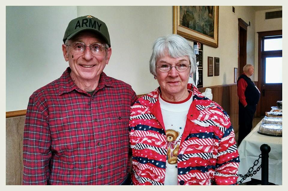 Man in red plaid shirt and Army hat stands with arm around woman in patriotic sweater and glasses