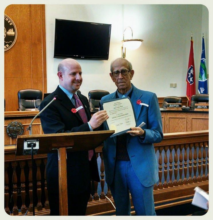 Man in black suit presents certificate to man in blue suit, both are standing at wooden podium at front of courtroom