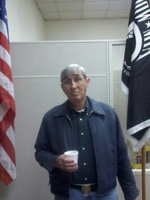 Man stands between American flag and POW-MIA flag, holding coffee cup and looking into camera