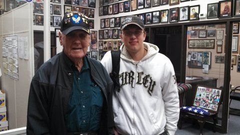 Man in Air Force hat and black jacket stands beside man in white sweatshirt and grey hat