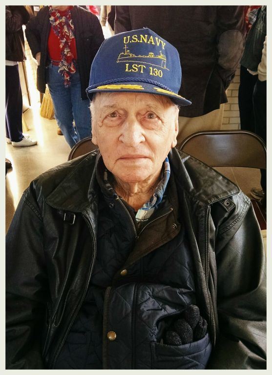 Up close picture of man wearing black leather jacket, and blue US Navy LST 130 hat, veteran is looking into camera