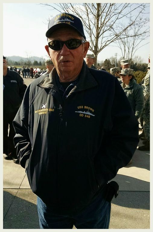 Man wearing black jacket and hat both with U.S.S. Brown, standing in parking lot hands in pockets, looking into camera