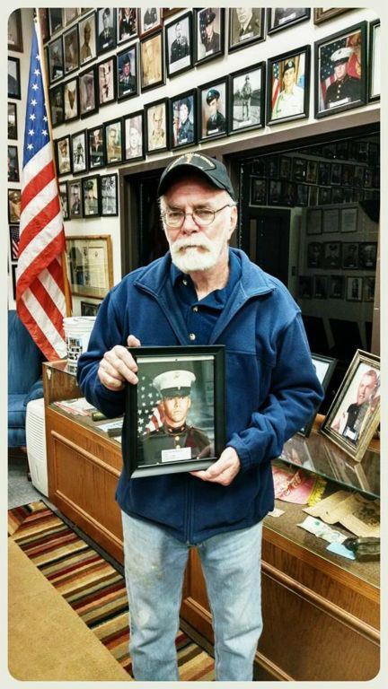 Man in blue stands in Veterans Affairs office, holding portrait of young marine