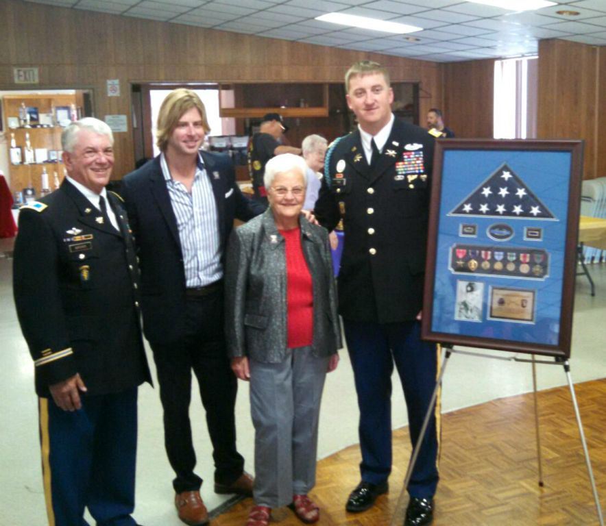 Two men in uniform flank a man in a suit and a woman in greay dress pants, beside them is a frame with American flag and medals and photograph of veteran