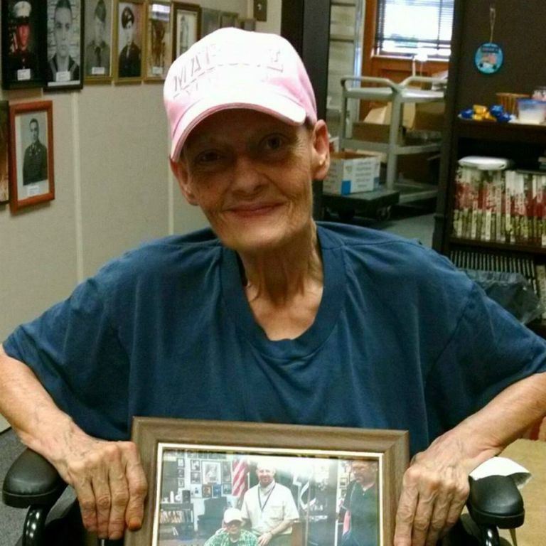 Woman in wheelchair, wearing blue shirt and pink hat, holds framed picture, she is looking into camera smiling