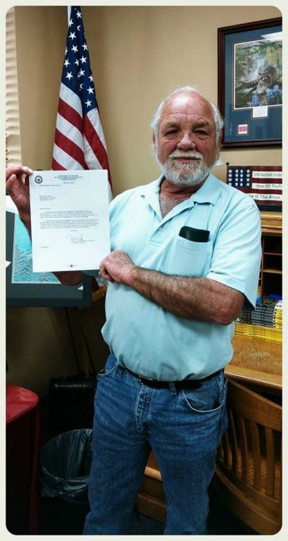 Man in blue shirt holds piece of formal paper up proudly, looking into camera smiling
